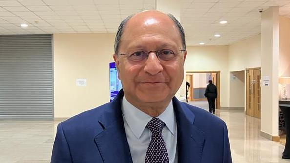 Shailesh Vara is standing in the middle of the image looking towards the camera with a light smile. He has short dark grey hair, and is wearing glasses and a white shirt, blue tie and suit jacket.