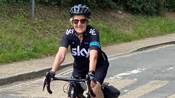 A woman with short grey hair rides a bike wearing sunglasses and a Sky branded cycling jersey and short. She is riding a bike down a road