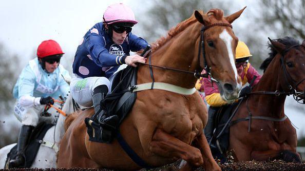 A close-up of a horse being ridden by a jockey in a blue shirt with pink polka dots and a pink cap, leaping over a jump with two other horses in hot pursuit