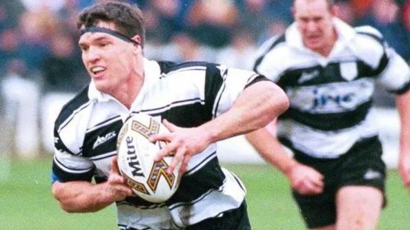 A man in a black and white striped Hull FC kit runs while grasping a rugby ball during a match. He has short dark hair and wears a head band and mouth guard. One of his team mates runs behind him.