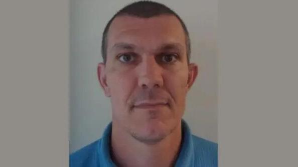 A custody image of Gary Rootham. He has very short, dark hair, and is looking directly into the camera against a pale backdrop. He is wearing a blue polo shirt and is unsmiling. 