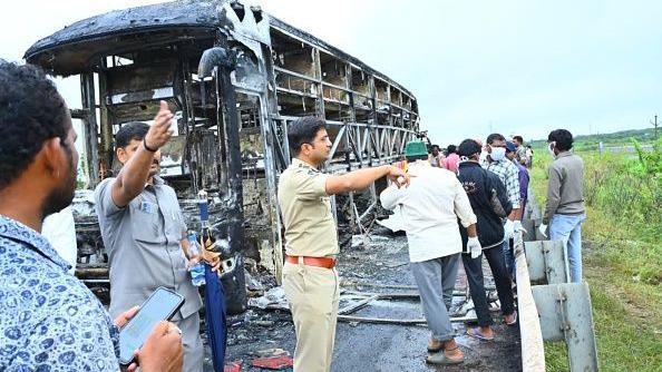 The charred remains of the bus that caught fire, with several people, including a police officer in uniform, standing nearby.