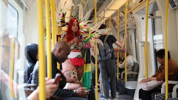 A woman with long read hair and wearing a feathered head dress, is looking at her phone while stood on a London Underground train. Other Tube passengers, not dressed in Carnival clothes, are seated around her.