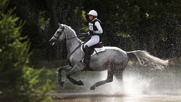 Photo of Georgie Campbell riding a grey horse on a cross country trial through water. The horse has three feet in the air and is clearly moving at speed. The water is spraying up as she rides through. 