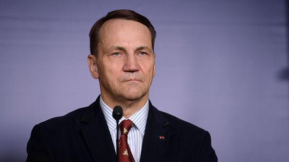 Polish Foreign Minister Radoslaw Sikorski in front of a microphone, wearing a jacket and red tie