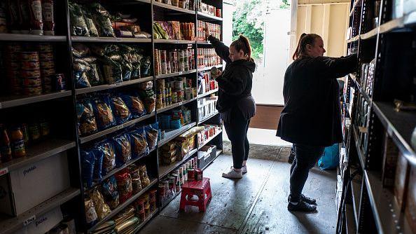 Two workers in a food bank organise supplies on shelves, which have supplies of tinned and dry food. There is a step on the floor to reach the higher shelves and a window behind the workers.