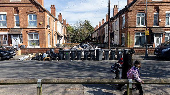 A woman and two children walk on the opposite side of the road to a row of black wheeled bins and rubbish left by the side of them. The bins are on the pavement near two rows of terraced houses in Bromfield Close, Aston, on 8 April.