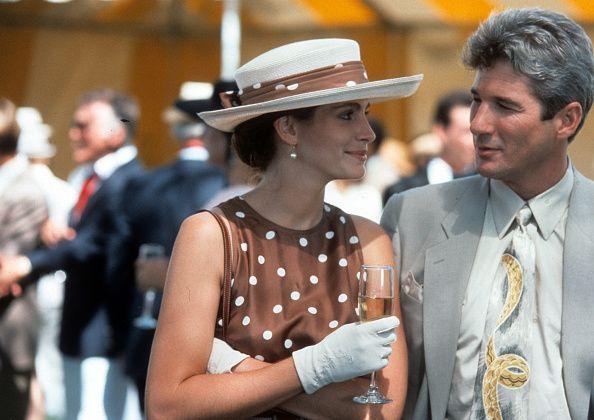 Julia Roberts has a drink with Richard Gere in a scene from the film 'Pretty Woman'. They are attending the polo and she wears a brown dress with white polka dots, a white straw hat, with a sash on it with the same material as her dress. She is turned towards Gere, smiling and is clutching champagne in one had and clutching herself in the other. Gere is looking back at her and is wearing a light grey shirt and blazer and a grey tie with a yellow snake deign on it. The background is blurred and show people at the polo event behind them