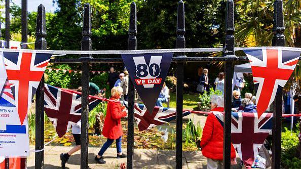 Flags and bunting are put up on railings for the 80th anniversary of VE Day. People have gathered in a park for a party on a sunny day.
