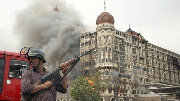 An Indian security official wearing a red striped shirt and a helmet is holding a gun as he stands alert as smoke and flames billow out from a section of The Taj Mahal hotel in Mumbai on November 29, 2008