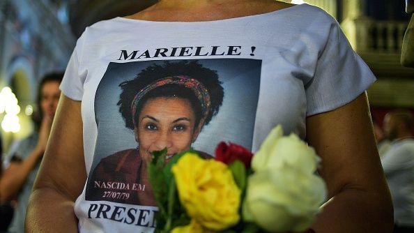 A woman wears a white t shirt with Marielle's face on it. She is also holding flowers. 