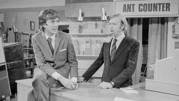 A black and white photograph of comedians Eric Idle (left) and Graham Chapman in the Buying an Ant sketch from the BBC television show Monty Python's Flying Circus in 1974.