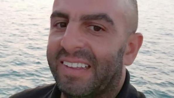 Samir, a man with very small hair, smiling for camera, with a water body in the background.