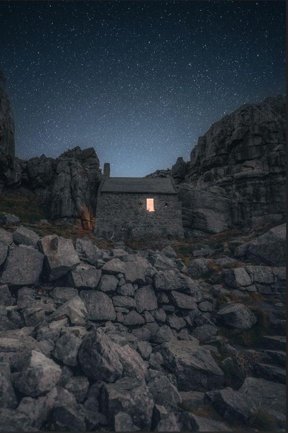 St Govan's Chapel is pictured against the backdrop of starry skies, with rocks in the foreground. The building is a simple stone rectangular structure with a small window, from which a light shines. The chapel is surrounded by stone cliffs.