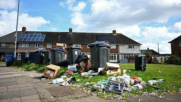 Waste spills from overflowing and uncollected wheelie bins outside homes in the Shard End suburb of Birmingham, on 14 April 2025. The rubbish strewn over an area of grass includes carboard boxes, a cutlery tray, household waste in bags and litter.