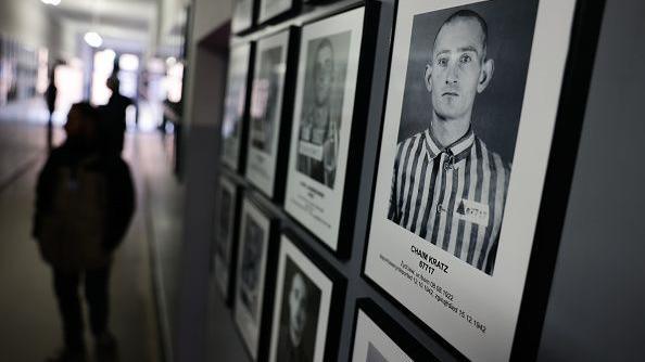 Black and white portraits of prisoners line a wall at the former Nazi German concentration camp Auschwitz-Brikenau. People visiting the site are walking down the corridor.