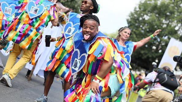 A man, with his tongue sticking out and wearing a multi-coloured costume, is pictured dancing with others in the background