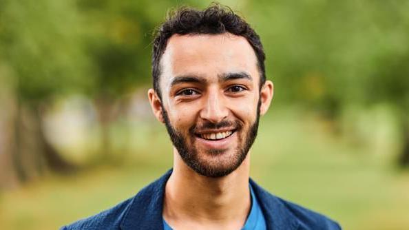 Dylan Walch smiles at the camera, He has dark hair and dark beard and is wearing a blue jacket and turquise T-shirt. There is a blurred background of grass and trees behind him.