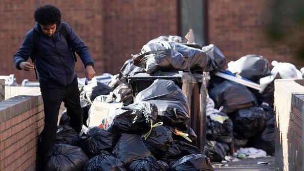A man picks his way past a large pile of rubbish on Bromfield Close in Aston on 8 April. Filled black bin bags are heaped up, blocking the path and there is also cardboard and wood on the pile.