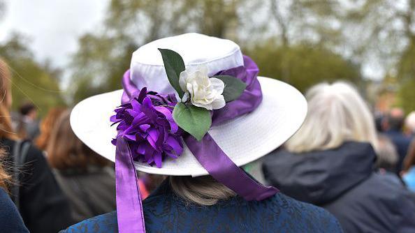 A woman wearing a hat decorated in the colours of the suffragette movement which fought for votes for women.