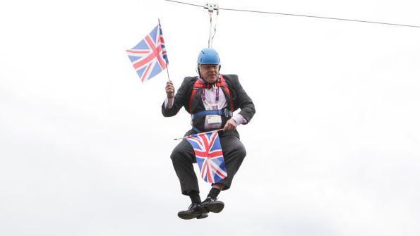 Boris Johnson dangles on a zipwire, seemingly stuck. He is wearing a helmet and has a Union Flag in either hand. 