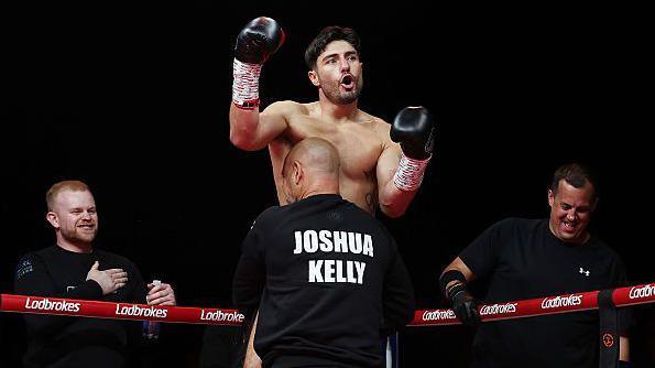 Josh Kelly is held up in the air by Adam Booth with two of his training team stood either side on the outside of the ropes of a boxing ring