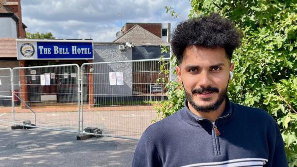 Wael, a man with curly hair, is wearing a navy blue jumper.  He stands in front of fences outside the Bell hotel in Epping which has temporary metal fences outside.