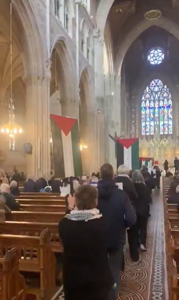 A place of people walked down the nave of a church with pews on either side. Some of the people are holding placards with Palestinian flags. In the background a large stained glass window.