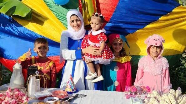 Woman standing with four children - all wearing colourful clothing - standing in front of rainbow coloured cloth and behind a table that is filled with party food