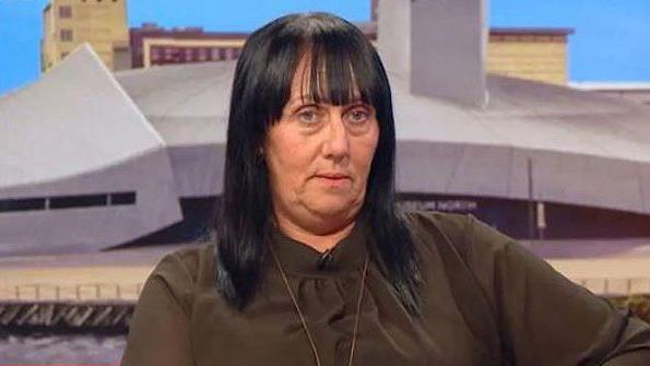 Debbie Duncan with long black hair wearing a brown blouse sitting on the sofa in BBC Breakfast TV's studio with Salford Quays in the background.  