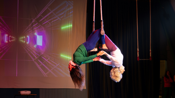 Two women hang upside down from a trapeze by their knees, holding hands as they face away from one another.