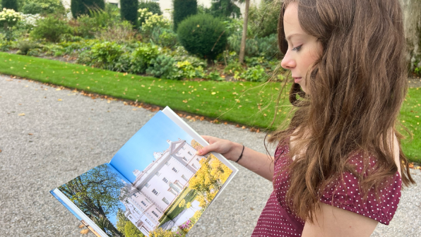 A woman looking at a book with a photograph of a large house in it. The woman has brunette hair and she is wearing a burgundy top.