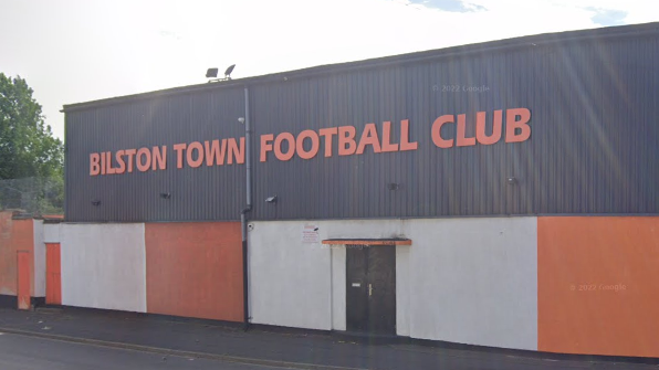 The club's orange and black or grey insignia is on the outside of a long, square building next to a main road with the name of the club in orange letters on the top exterior.