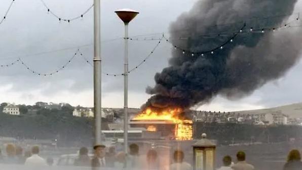 An archive picture of the Summerland complex on fire. A large column of thick black smoke bellows up into the sky above the structure, which is glowing orange and yellow as it is engulfed in flames.