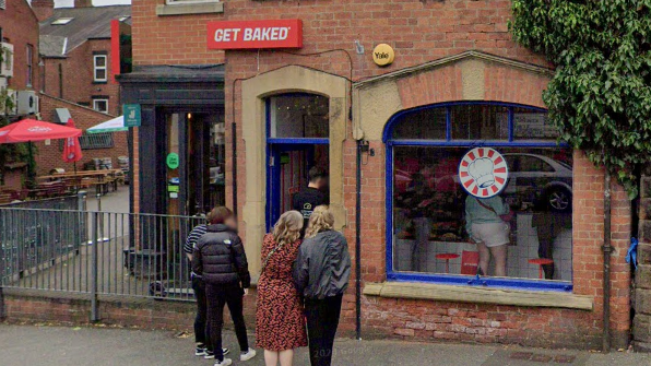 Customers stand outside a red brick building with a sign above the door reading Get Baked.