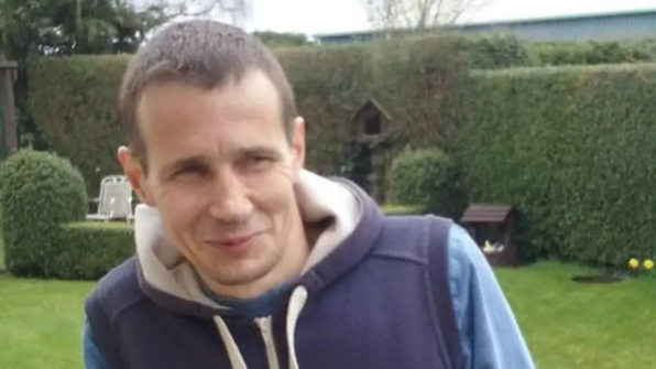 Neil Charles sits in a garden smiling and looking away from the camera. He wears a blue gilet jacket with a blue top underneath. He has short dark hair.