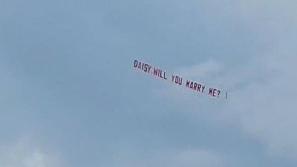 A sky banner being trailed by a plane reads 'Daisy will you marry me?' on a blue sky backdrop