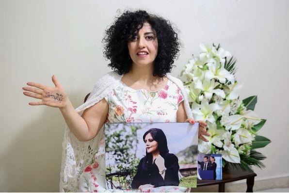 Narges Mohammadi, wearing a white dress with flower patterns on it, holds out her right hand to show a message written on it, while holding a photo in her left hand showing Mahsa Amini