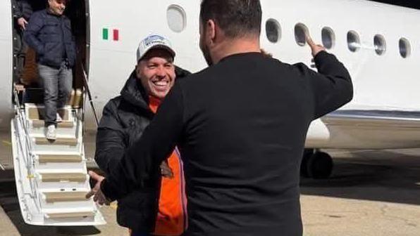 A man embraces another man after getting off a small passenger jet. The man facing the camera is smiling. He's wearing a black jacket, orange t-shirt and black jacket.