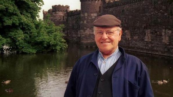 Fred Dibnah is standing in front of Beaumaris Castle on the island of Anglesey. The castle is surrounded by a moat. Dibnah is smiling at the camera and is wearing a brown flat cap along with a blue shirt, navy cardigan and blue coat.