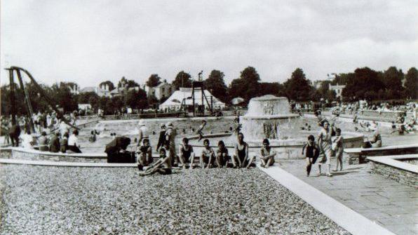 This black-and-white image captures Cheltenham Lido in 1981. A slide is visible to the left, while the poolside is bustling with activity, with children in swimsuits relaxing on the surrounding grass.