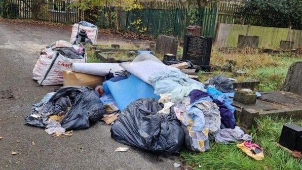 A pile of rubbish is strewn over several graves that have dark or black-coloured headstones. The piles include black bin bags, clothing, cardboard boxes and furniture.