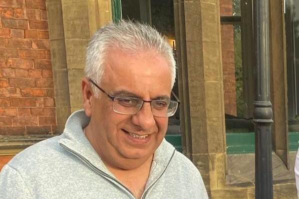 A photo of Paresh Thakkar, a man with white hair wearing black glasses and a grey zip-up sweatshirt. He smiles, standing in front of the hotel.