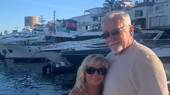 A woman with long blonde hair and wearing sunglasses and a sundress standing with a man who has short grey hair and a grey beard and is wearing a beige t-shirt. They are stood on a harbour with a number of yachts moored in the background.