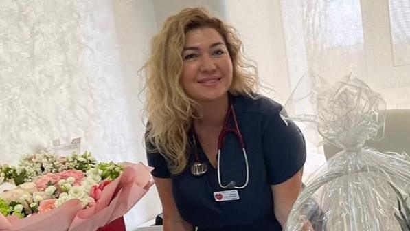 Dr Olga Pantas in medical scrubs next to a large bunch of flowers and fruit basket. She smiles into the camera and has long blonde hair with a stethoscope round her shoulders.