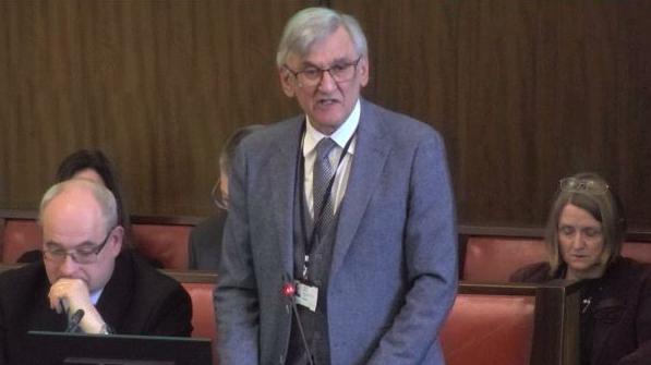 Conservative Councillor Peter Butlin, Warwickshire County Council's Portfolio Holder for Finance, stands to speak in the council chamber, There are red leather and wooden benches behind him and he is wearing a grey suit, tie and waistcoat.