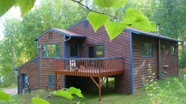 A wooden house with a deck on stilts. It is surrounded by hedges and trees. A small path leads away from the house.