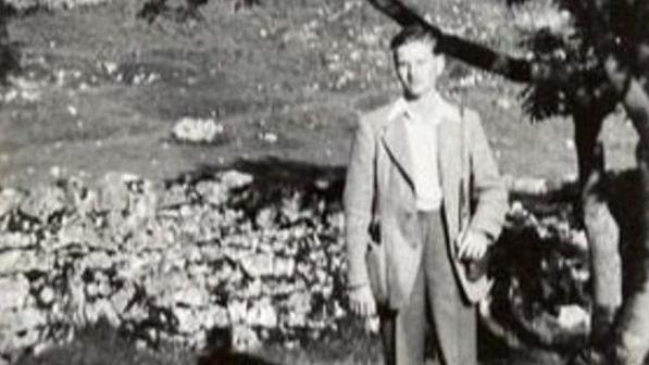 Gordon, aged 15, in slacks and a light jacket with a shirt underneath. He is outside standing on a wall next to a tree.