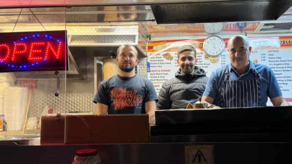 Three men standing behind a kebab shop counter with menus behind them and a large lit-up open sign.