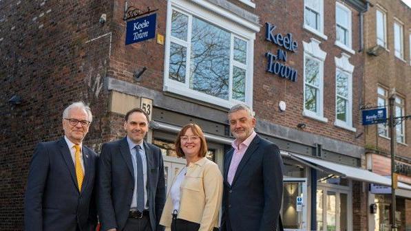 Four people, from the left, two men, a woman, then a man, standing outside a red brick building with the sign "Keele in Town". They are all wearing business wear and smiling towards the camera. 